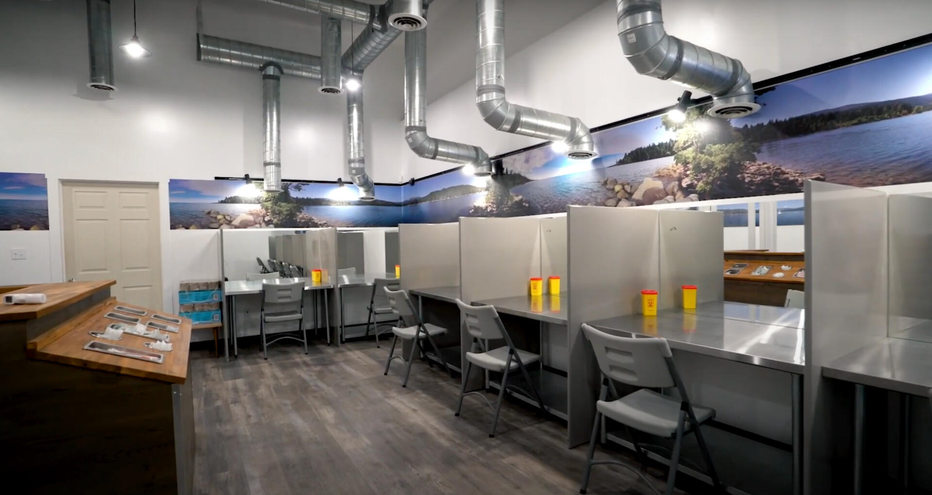 A large room with individual stalls, each containing a chair, a table, a sharps container, and an overhead fan.